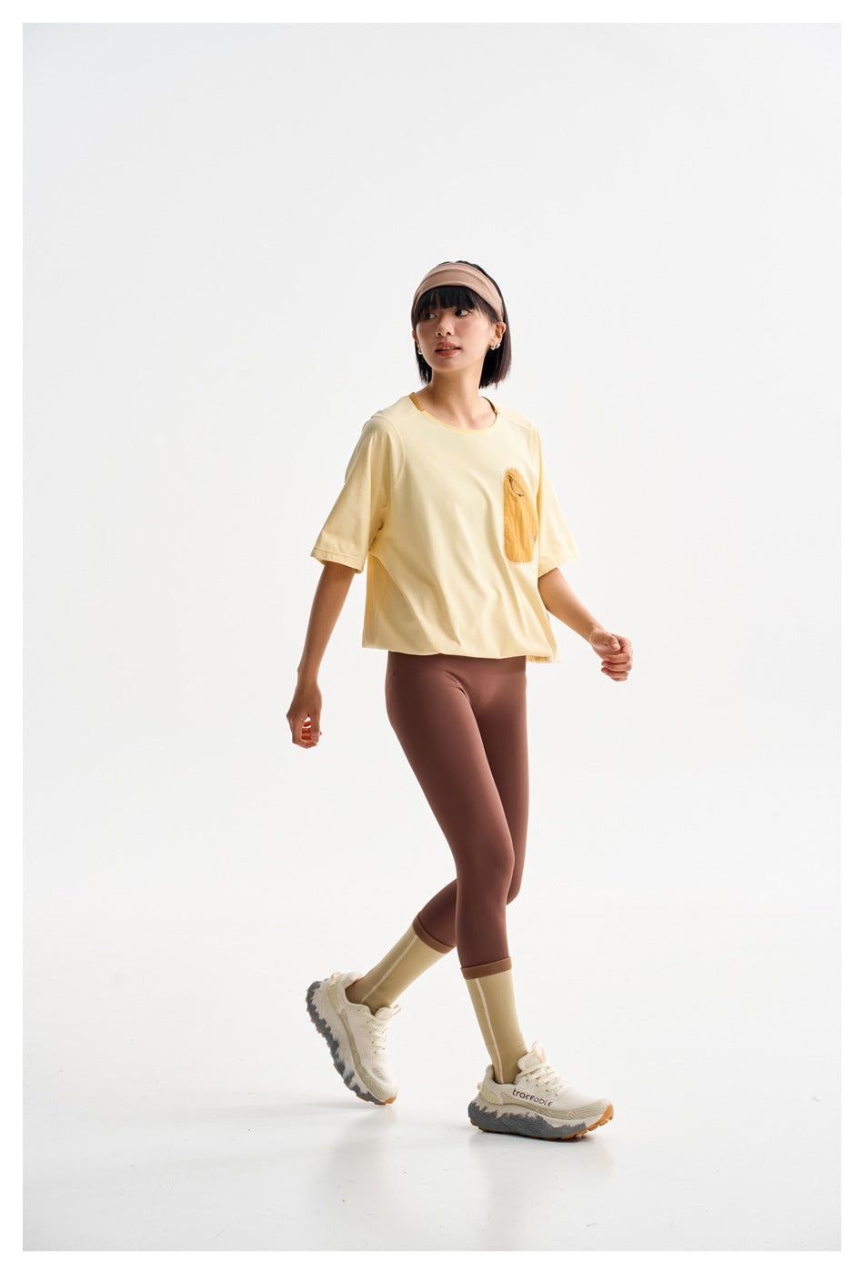 Model walking in a pale yellow oversized T-shirt with an ochre chest pocket and brown leggings.