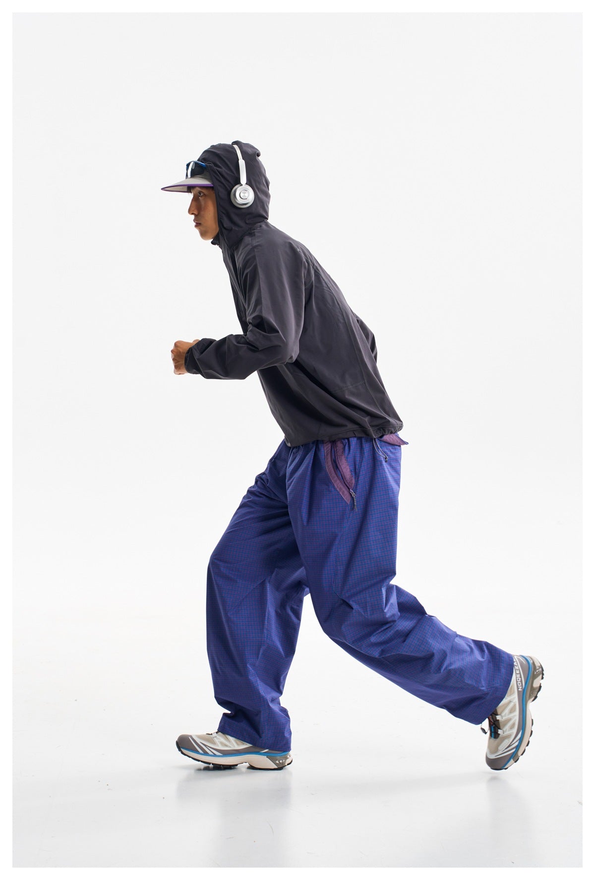 Side view of a running model wearing a dark hooded jacket and loose blue-purple checked trousers.