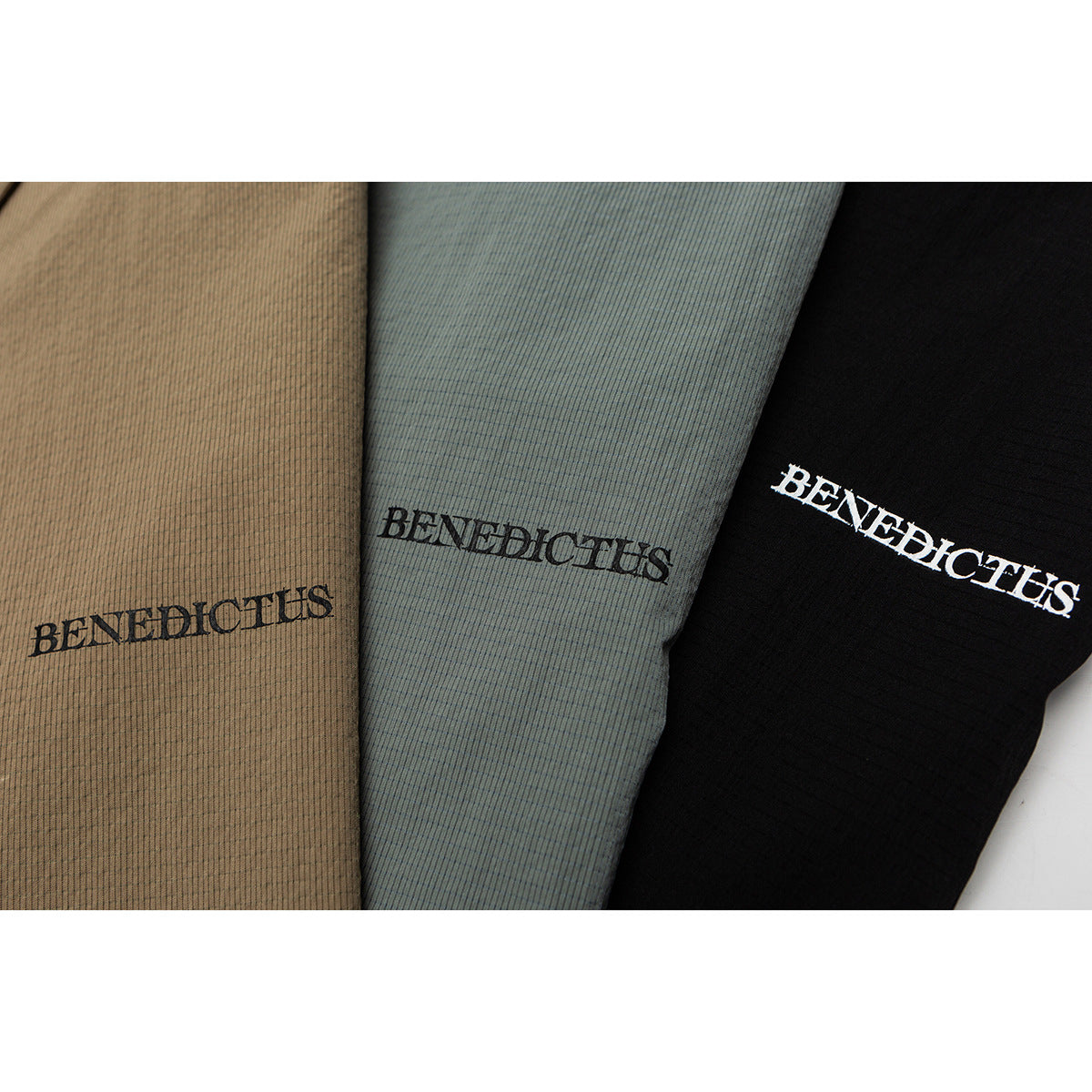 Close-up of tan, gray-green, and black shirts with BENEDICTUS lettering on the fabric.