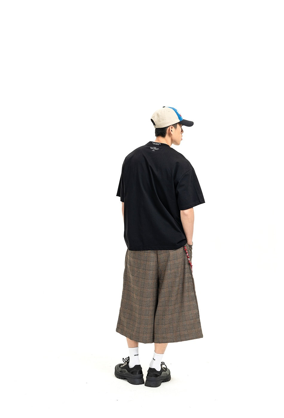 Rear view of a model wearing brown-gray plaid wide-leg shorts with a black T-shirt and sneakers.