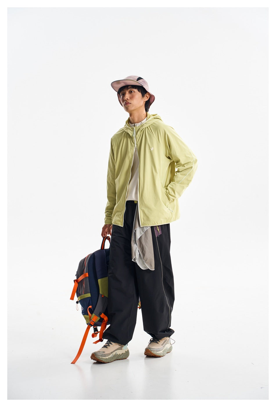 Model wearing a pale yellow-green hooded zip jacket and black trousers, carrying a navy backpack with orange trim.