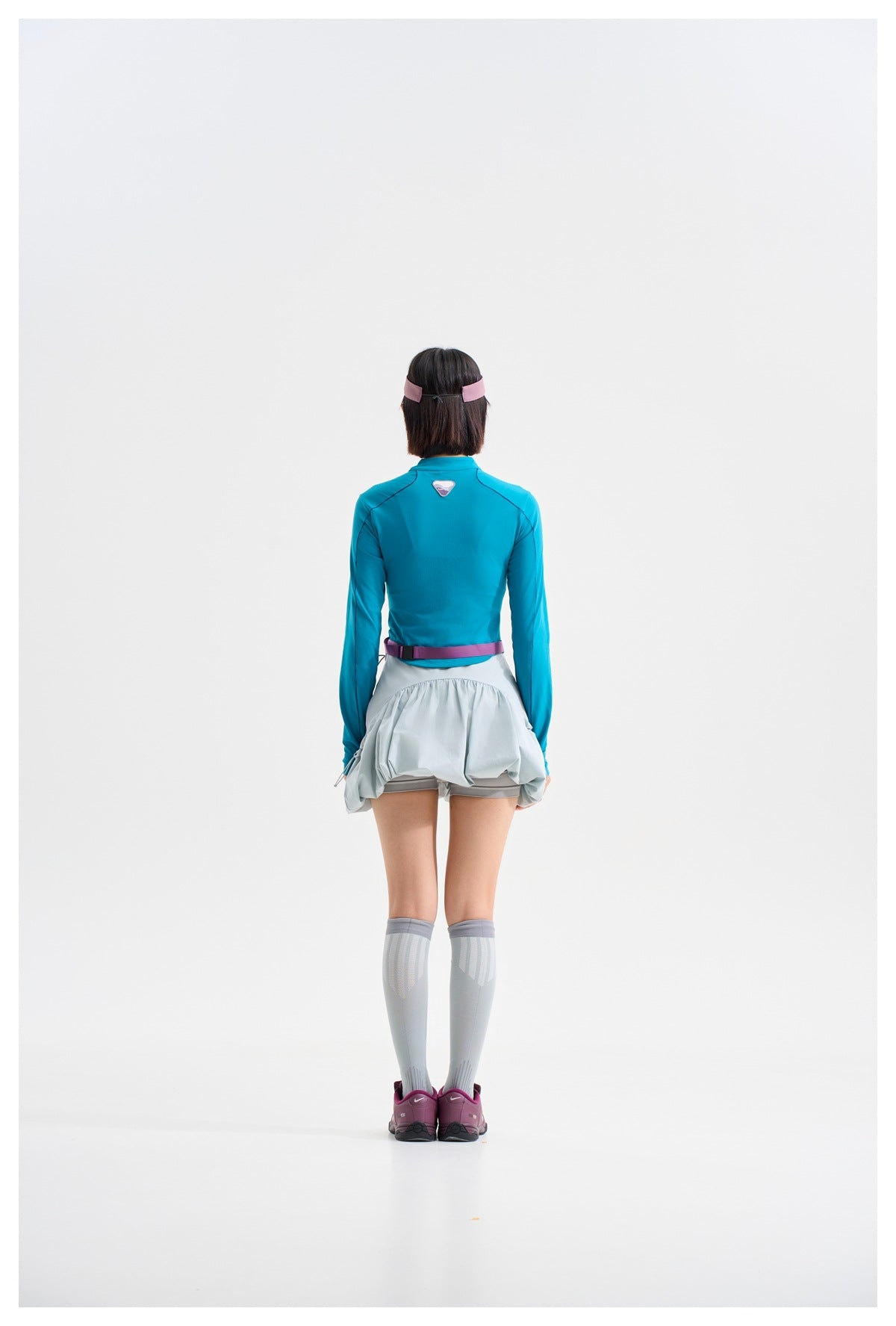 Rear view of a model wearing a teal long-sleeve top and pale blue-gray gathered skirt with a purple waistband.