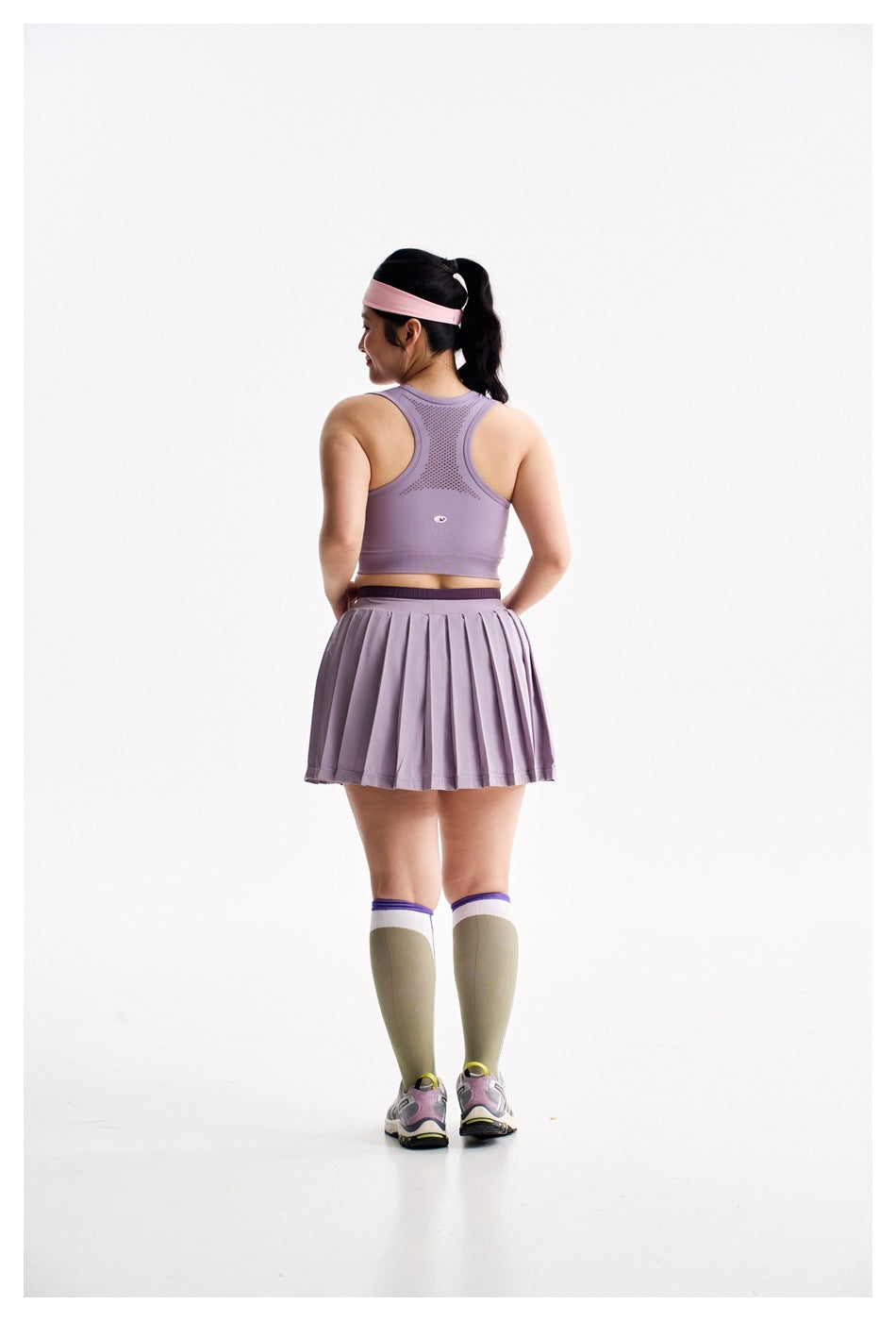 Full rear view of a model in a lavender racerback sports bra and pleated skirt, with knee-high socks and sneakers.