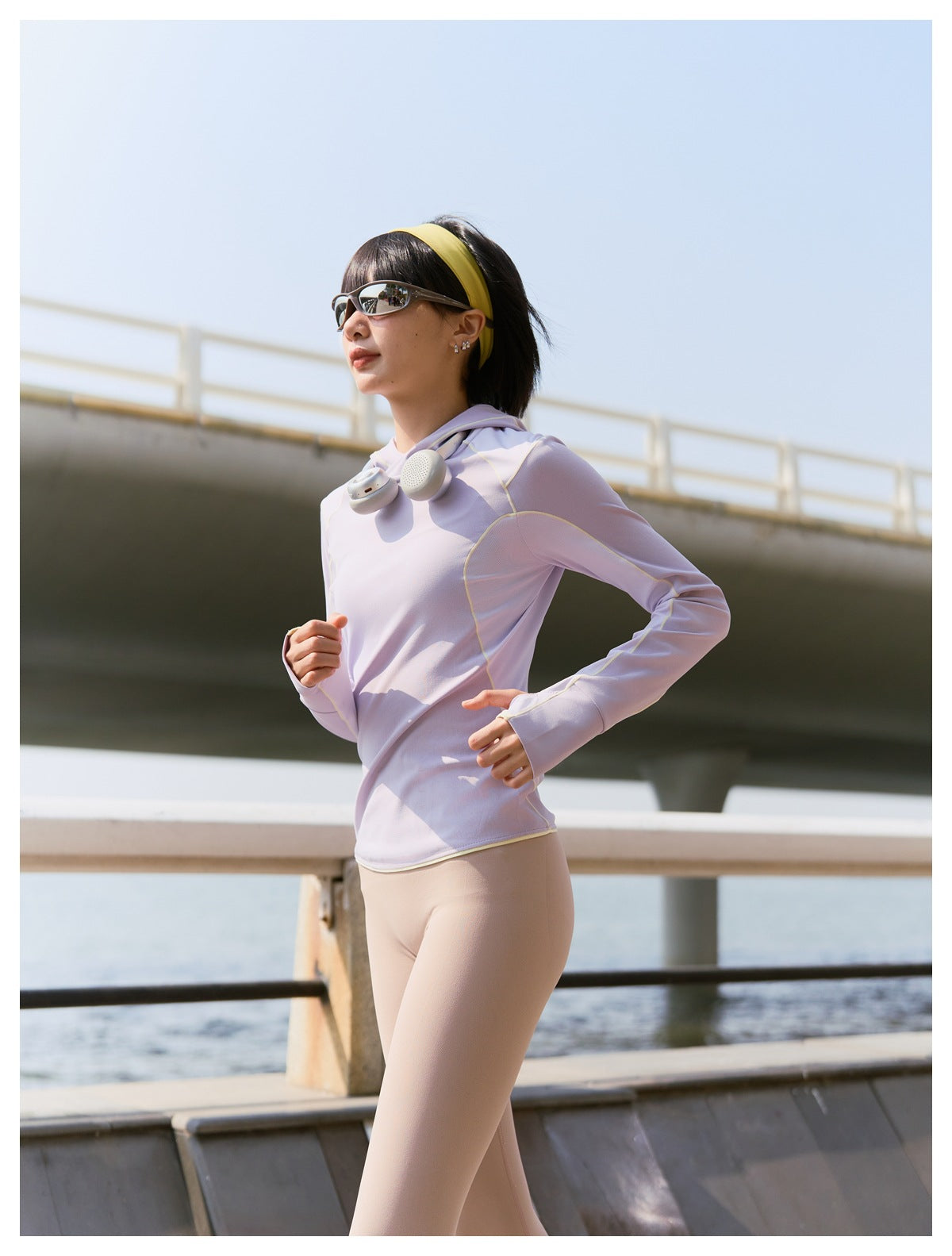 Model jogging outdoors in a lavender hooded athletic top with pale yellow piping and light-colored leggings.
