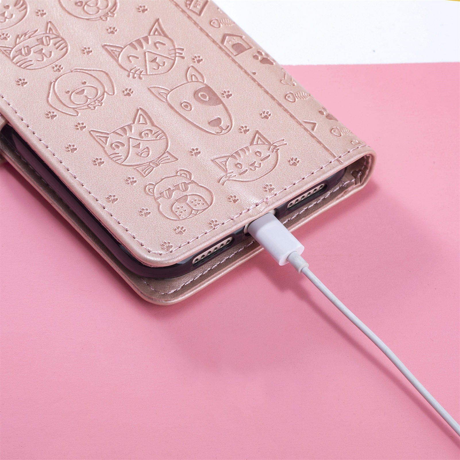 Close-up of an iPhone 16 Pro Max Wallet Case with Cute Cat and Dog Embossed Design, resting on a pink and white surface. A white charging cable is plugged into the phone.