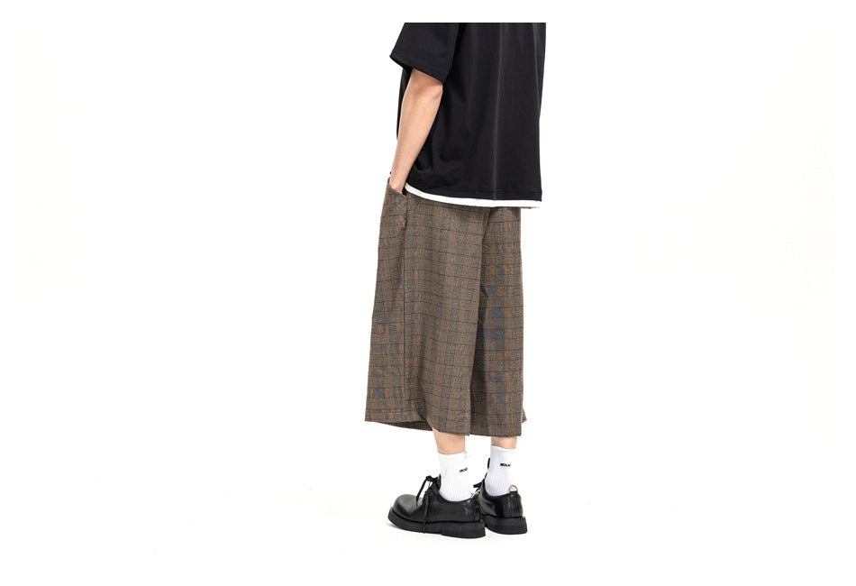 Rear three-quarter view of a model wearing brown plaid wide-leg shorts with a black T-shirt, white socks, and black shoes.