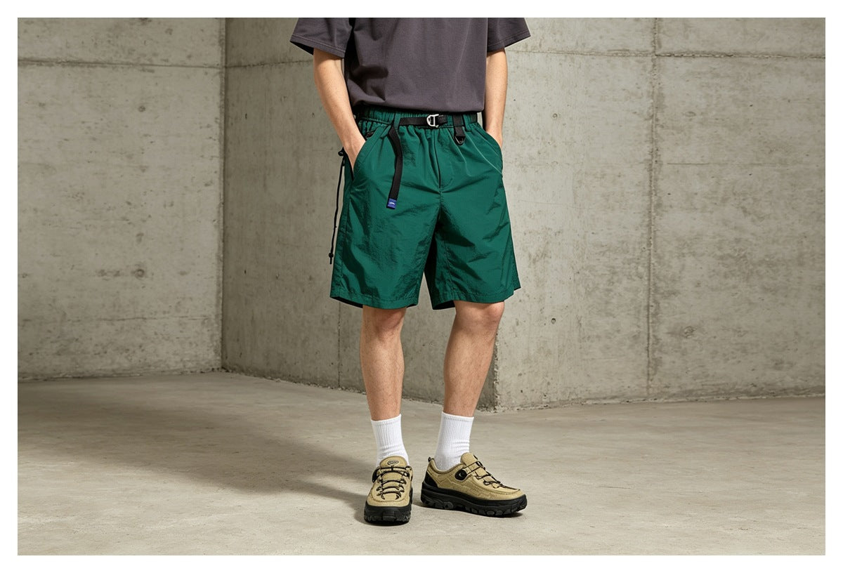 Cropped view of a model wearing green belted shorts with hands in pockets, gray tee and sneakers in a concrete setting