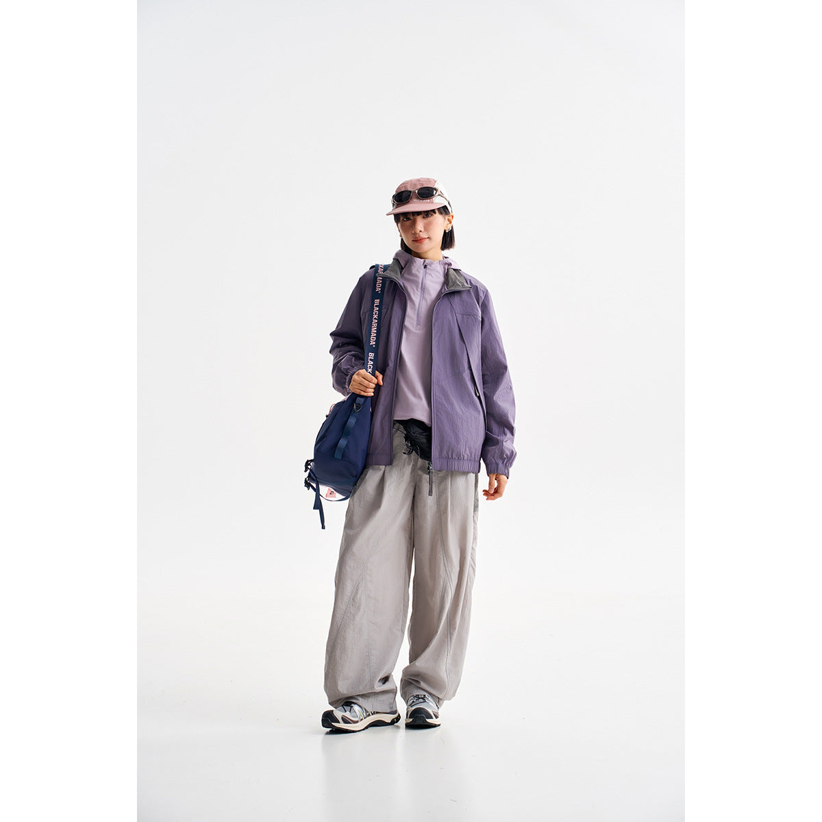 A model wears a purple zip jacket over a lavender top with loose gray trousers and a pink cap.