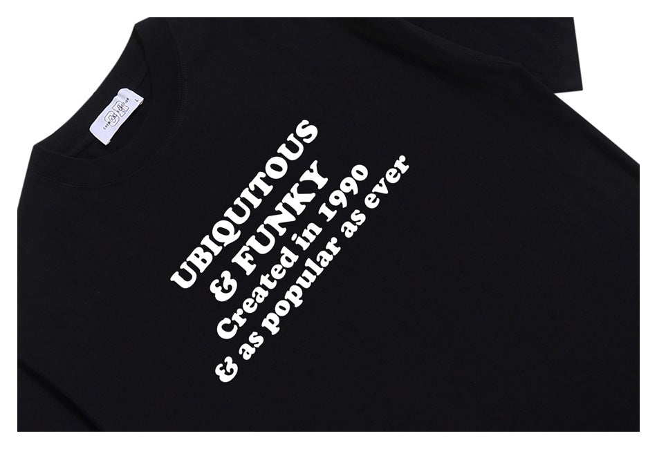 Angled flat lay of a black T-shirt with bold white text reading “UBIQUITOUS & FUNNY Created in 1990 & as popular as ever.”