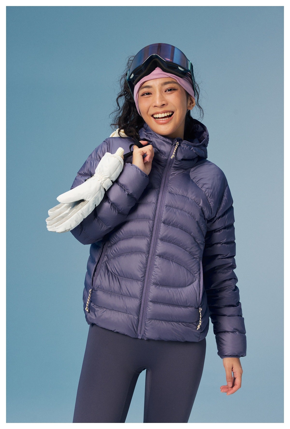 A smiling woman models a lavender hooded puffer jacket and leggings, holding white gloves.