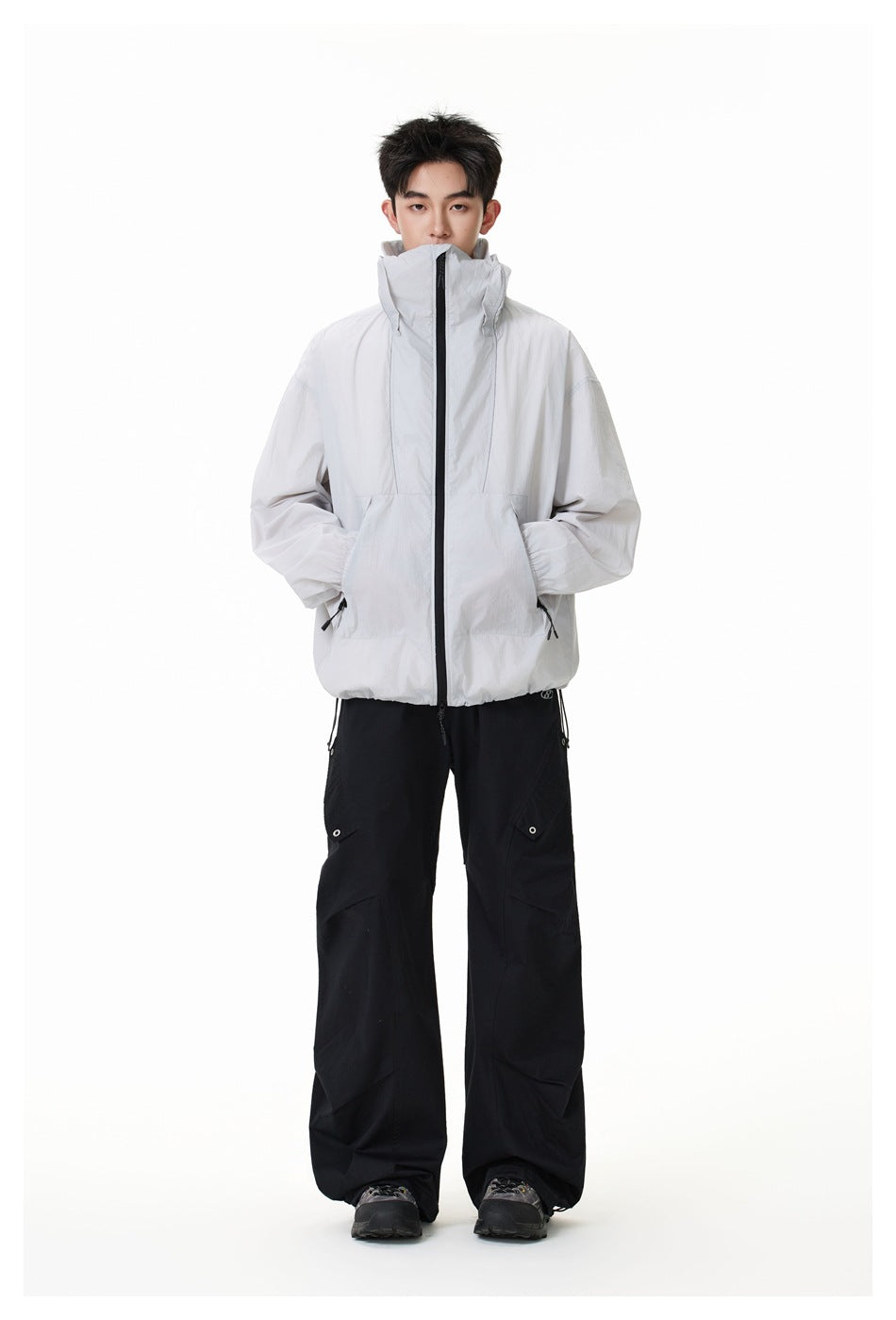 Full-length front view of a model in a light gray high-collar hooded jacket and loose black pants.