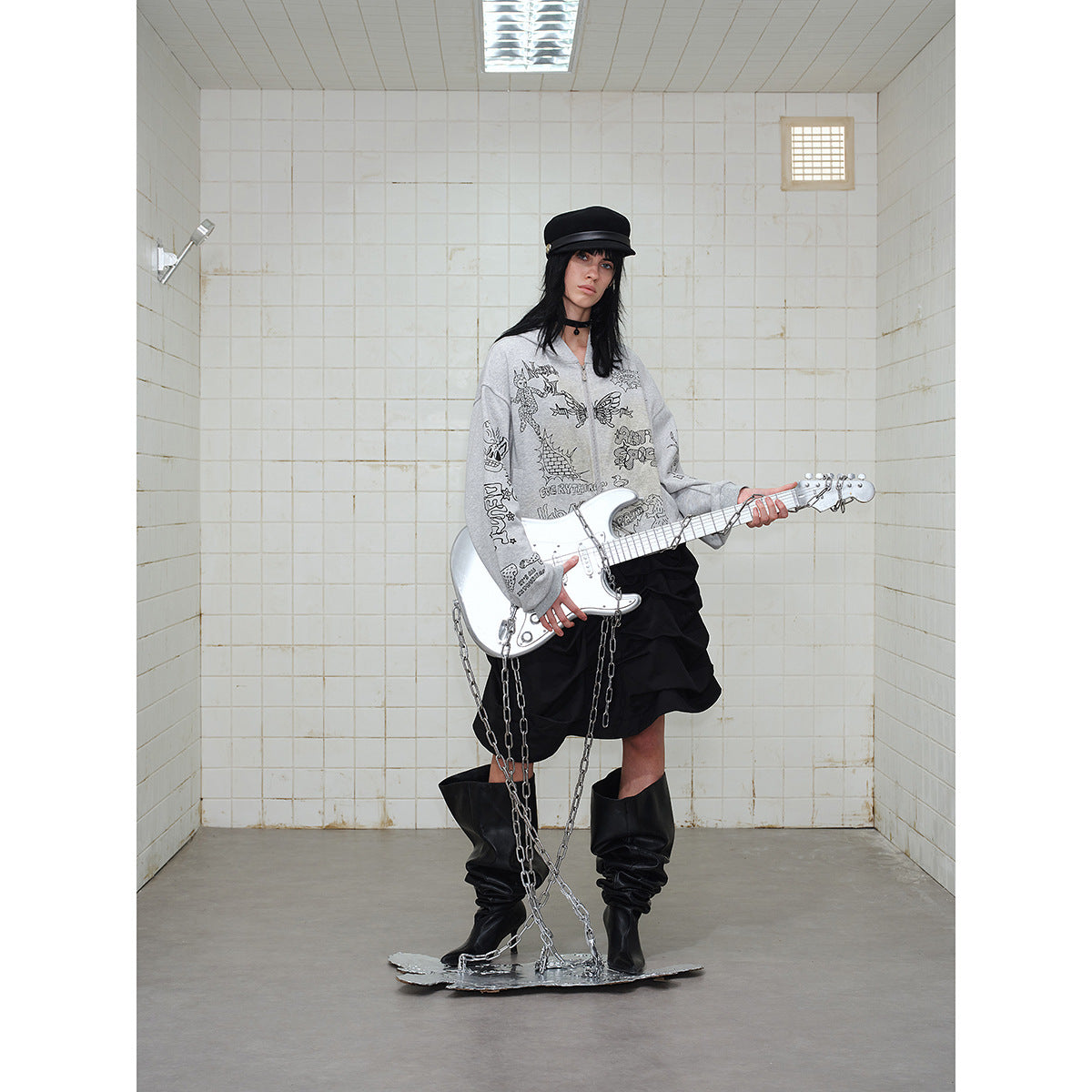 Model wearing a gray graphic zip-up hoodie and black ruffled skirt, holding a silver guitar.