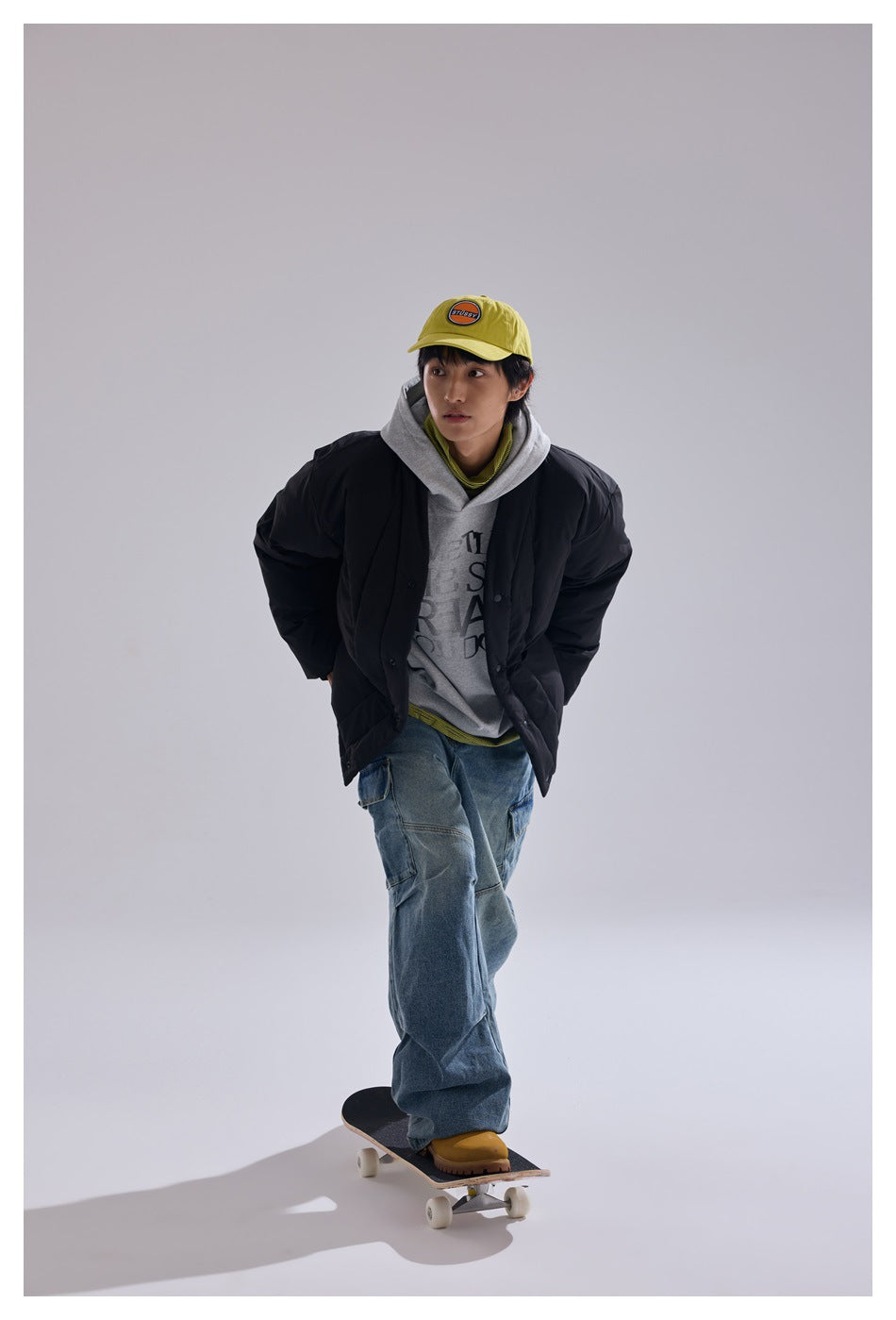 Full-length model wearing a black quilted jacket and gray hoodie while standing on a skateboard.