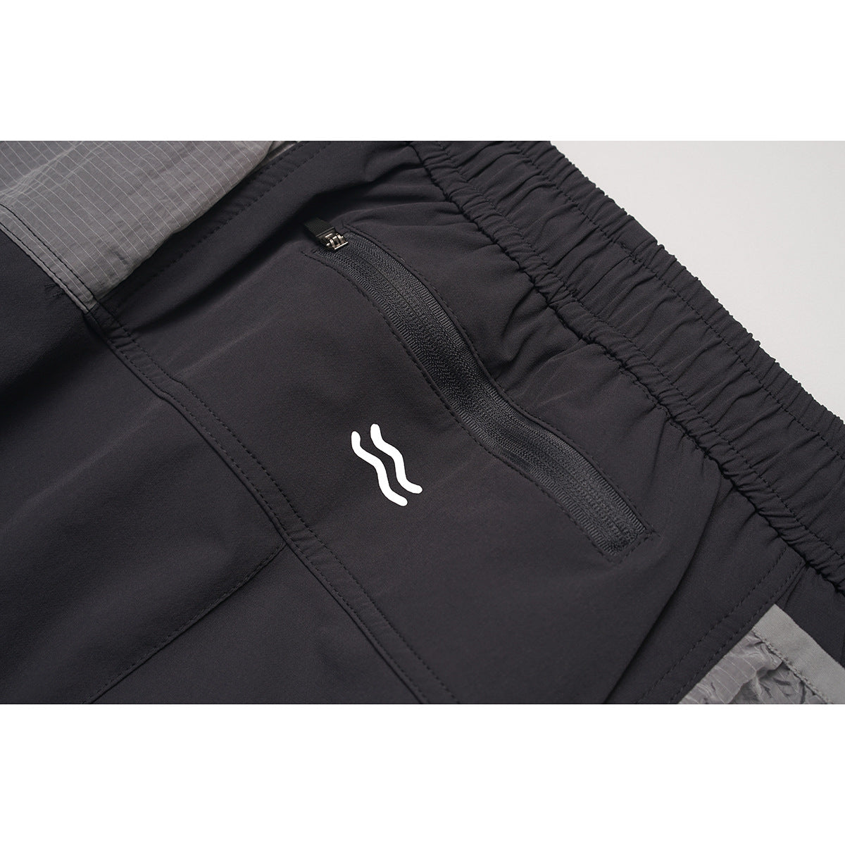 Close-up of charcoal pants with a zip pocket, small white logo, elastic waistband, and gray patterned panel.