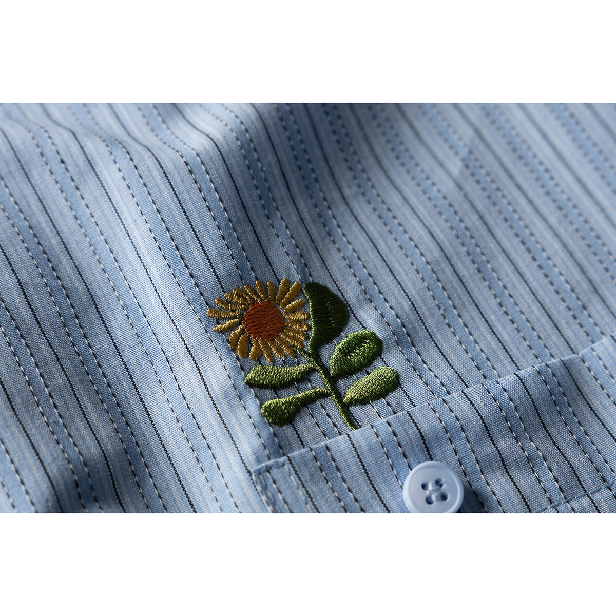 Embroidered yellow flower with green leaves above a pocket on a pale blue striped shirt.