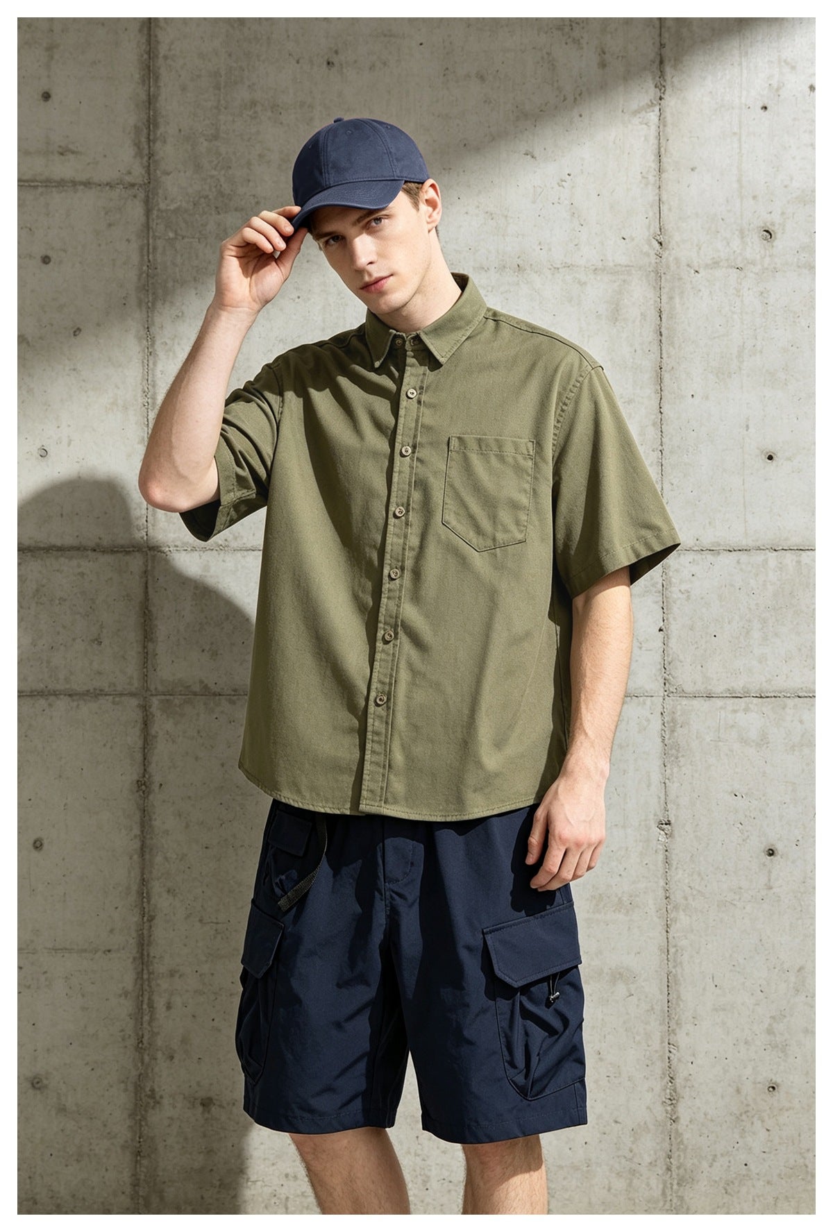 Man in army green short-sleeve button-up shirt touching his navy cap, with navy cargo shorts, in front of concrete