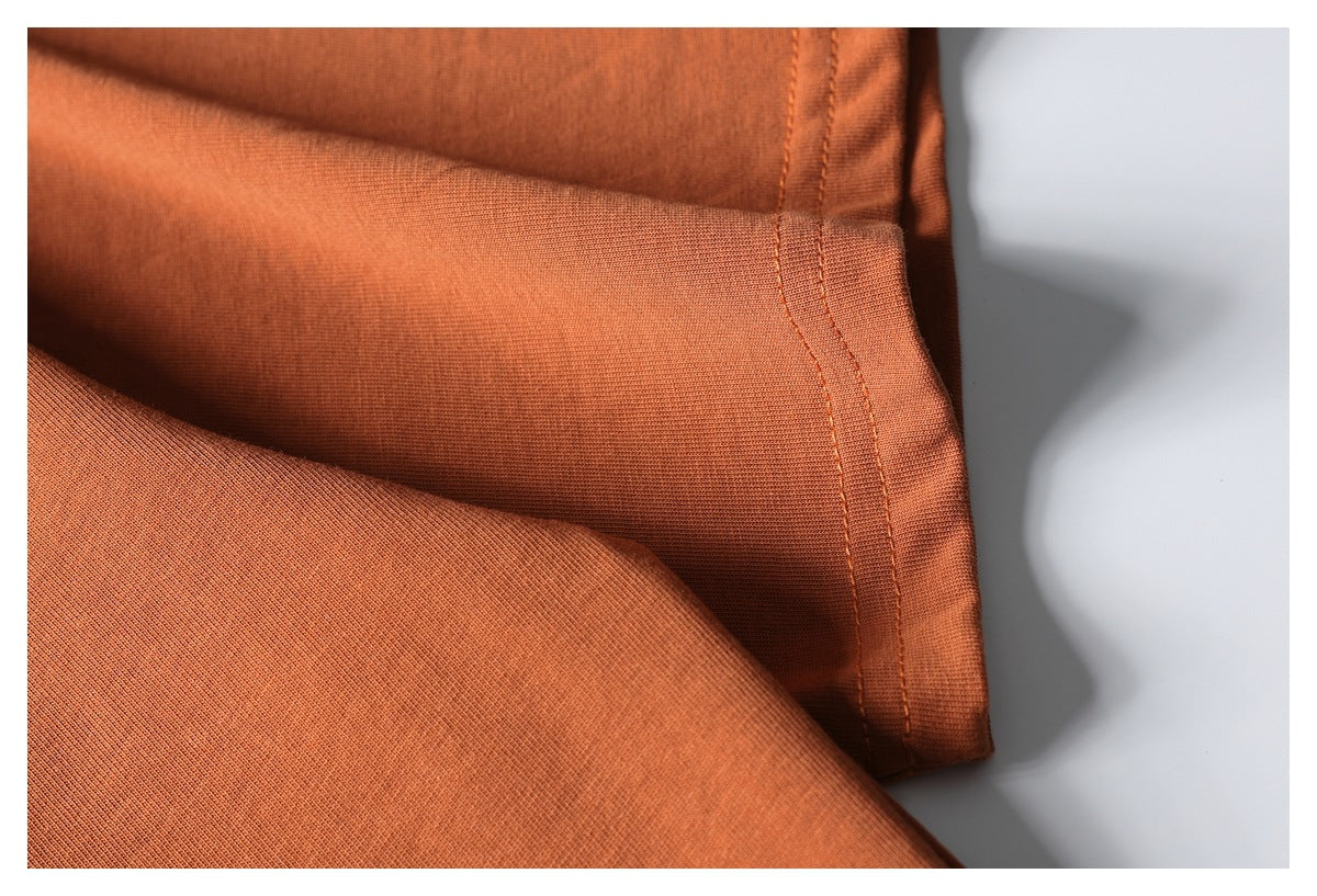 Close-up of rust-orange T-shirt fabric and a stitched side edge, laid flat against a pale background.