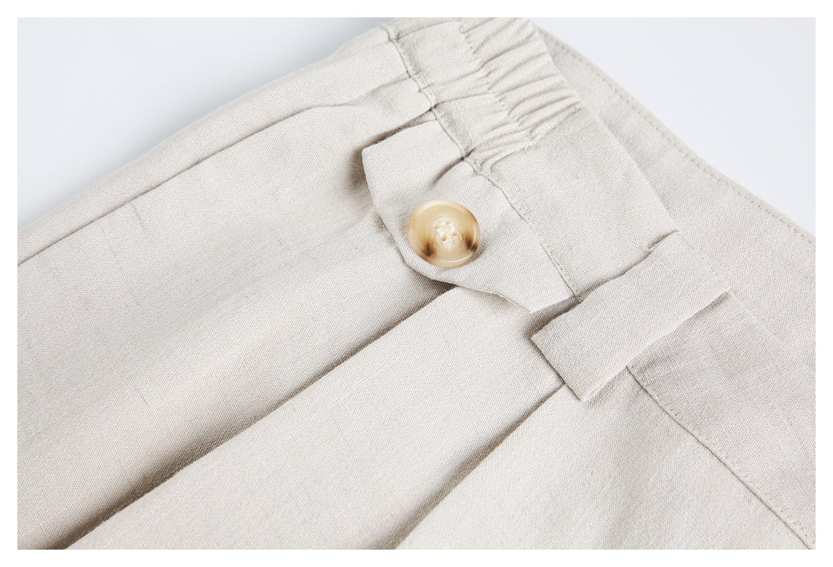 Close-up of ivory trousers showing a tan button closure, belt loops, pleats, and gathered elastic at the waist.