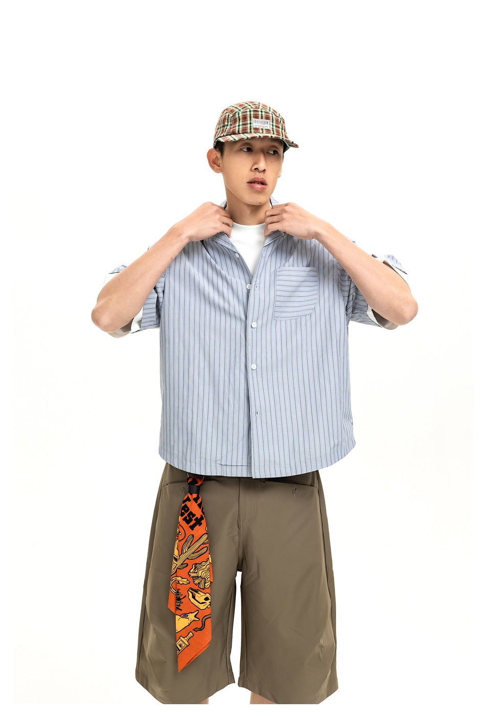 Front view of a model wearing a light blue striped button-up shirt, olive shorts, plaid cap and orange patterned scarf.