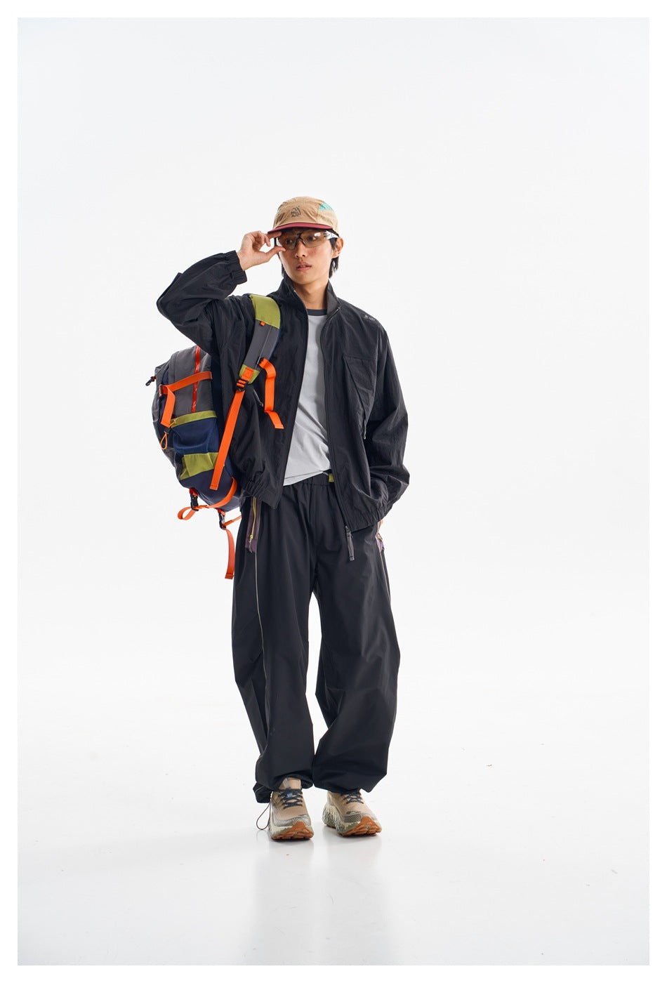 Full-length model in a black jacket and loose trousers, wearing a cap and carrying a navy backpack with orange straps.