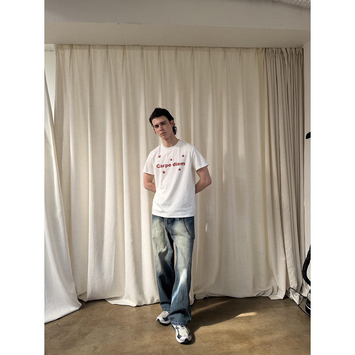 Model wearing a white Carpe diem star-print T-shirt and loose faded jeans, posing with hands behind his back.