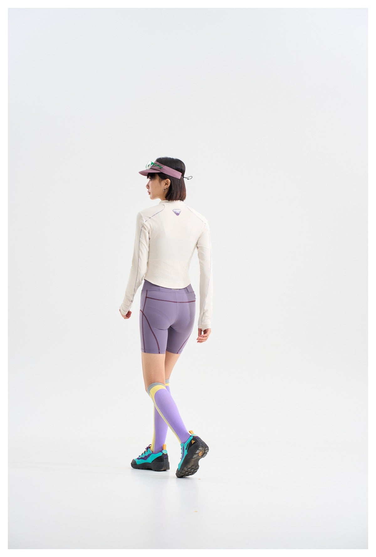 Rear view of a model wearing a white long-sleeve top, lavender cycling shorts, and color-block knee-high socks.