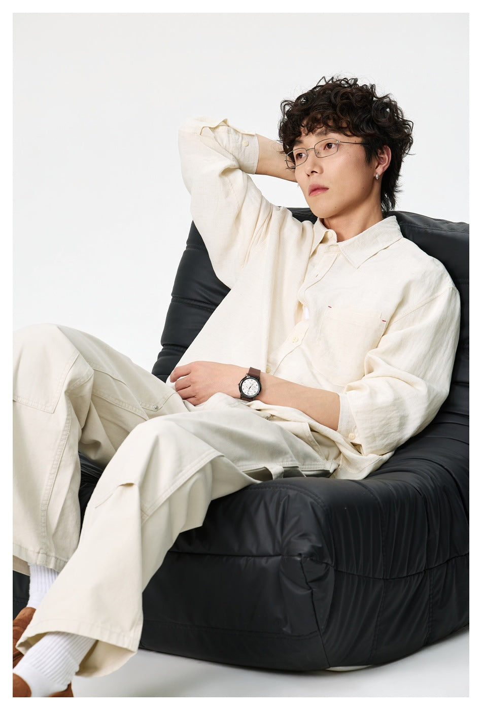 Man seated on a black chair wearing a cream button-up shirt with chest pocket and light pants
