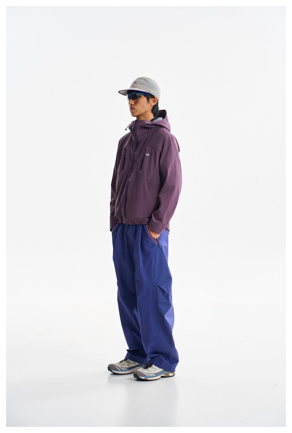 Three-quarter view of a model wearing a plum hooded zip jacket and loose blue-purple checked trousers.