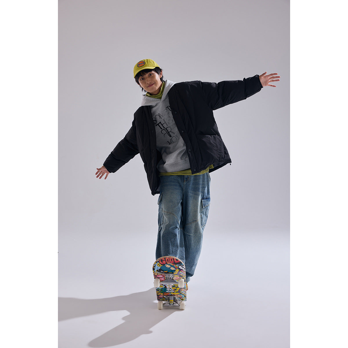Model wearing a black oversized quilted jacket over a gray hoodie, shown full-length with a skateboard.