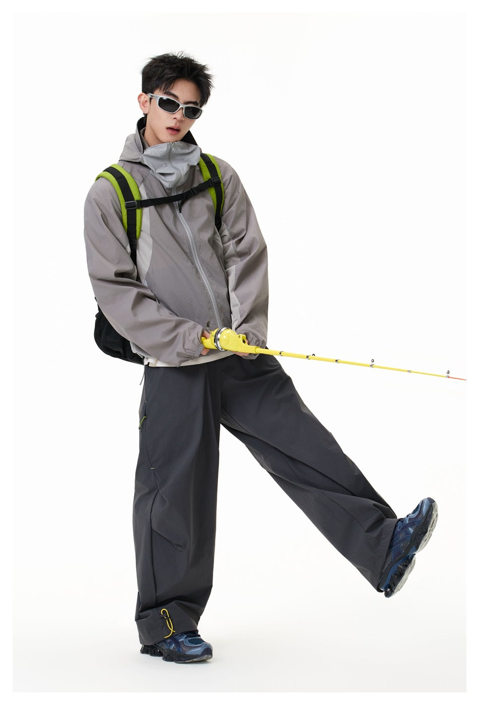 Model wearing the gray hooded zip jacket with gray collar panel, dark pants and a green backpack, holding a yellow fishing