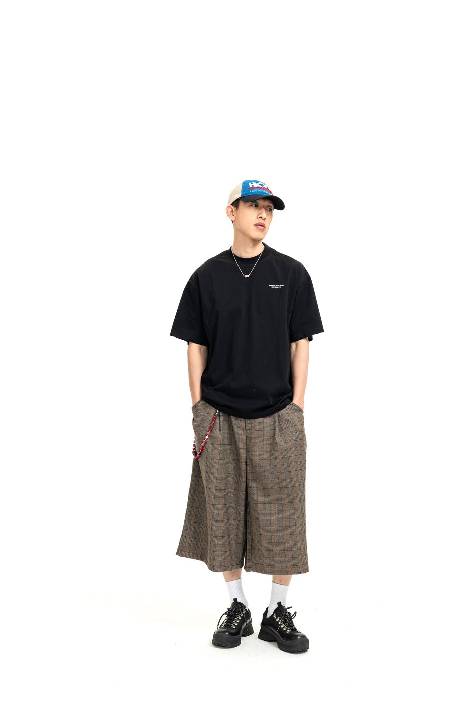 Full-length model wearing a loose black T-shirt, brown plaid wide-leg shorts, and a color-block cap.