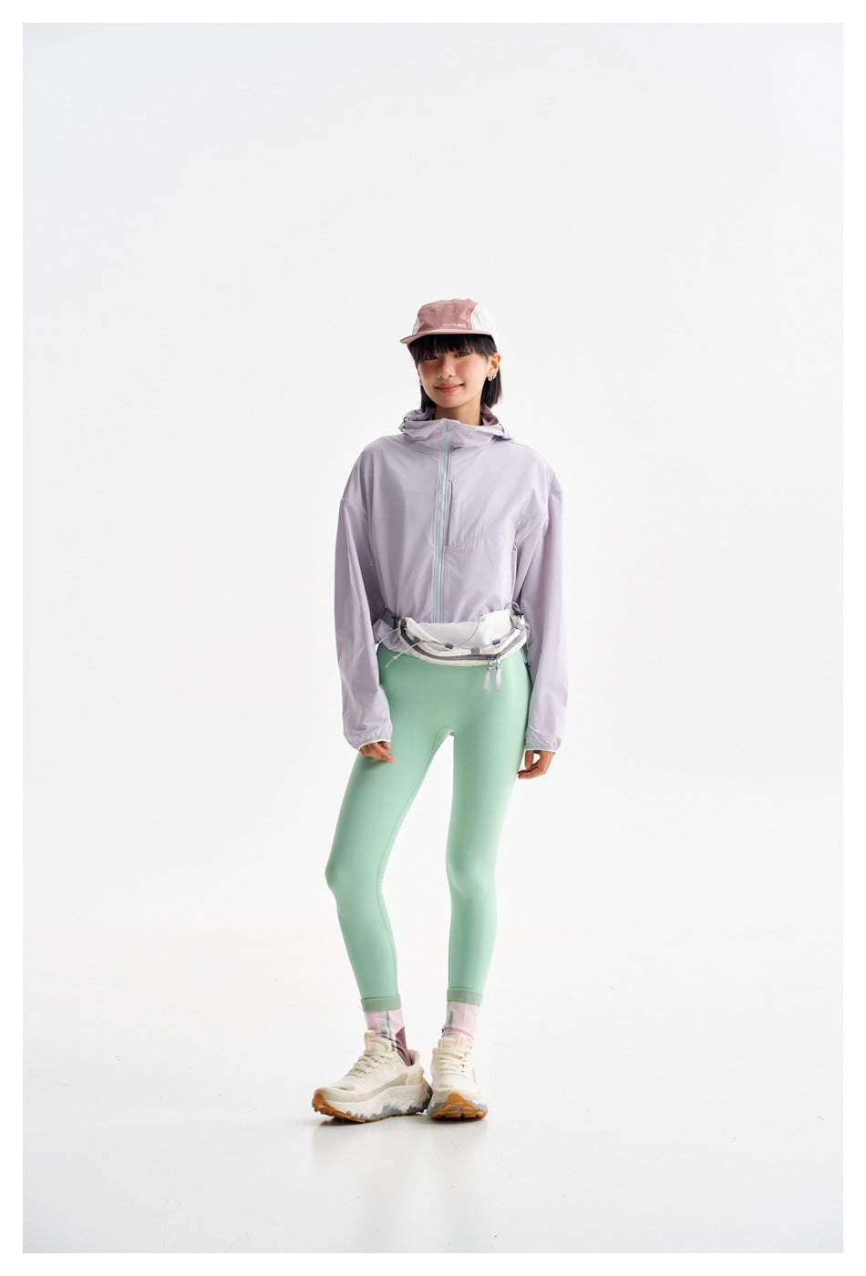 Model wearing a pale lavender zip-front hooded jacket, mint leggings, and a white waist bag.