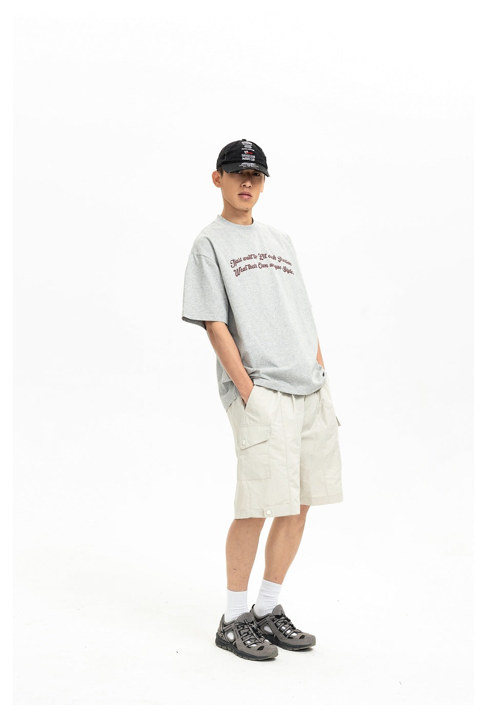 Full-length model wearing a light gray graphic T-shirt, cream cargo shorts, a black cap, and gray sneakers.