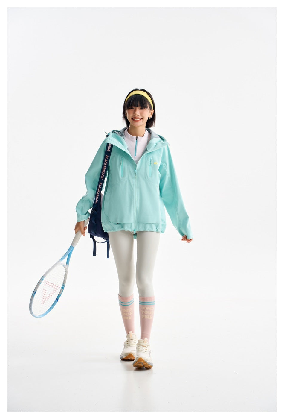 Model wearing a mint hooded zip jacket with chest pockets, white leggings and pink socks, holding a tennis racket.