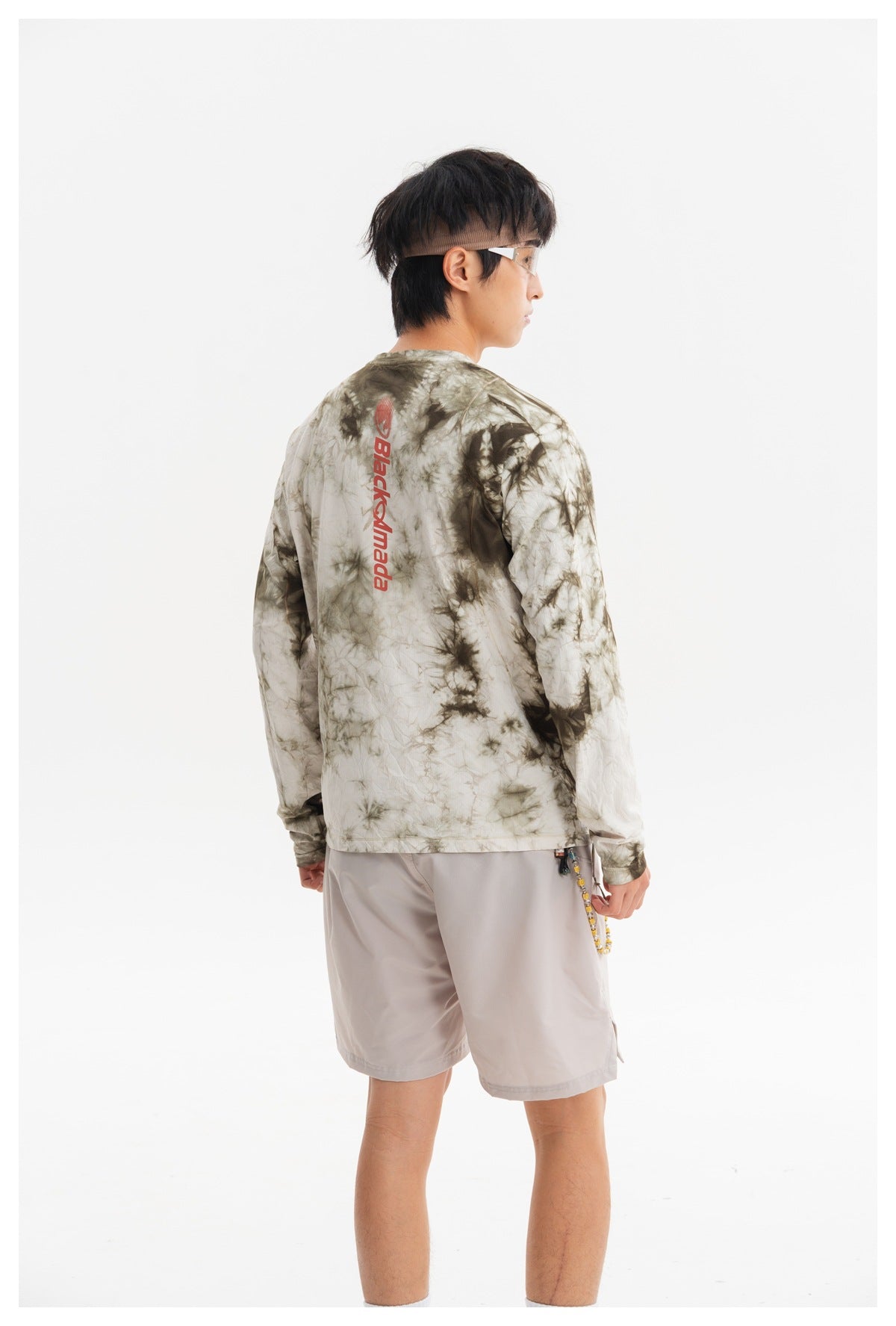 Rear view of a model wearing an olive-gray tie-dye long-sleeve shirt with a vertical red back print and pale gray shorts.