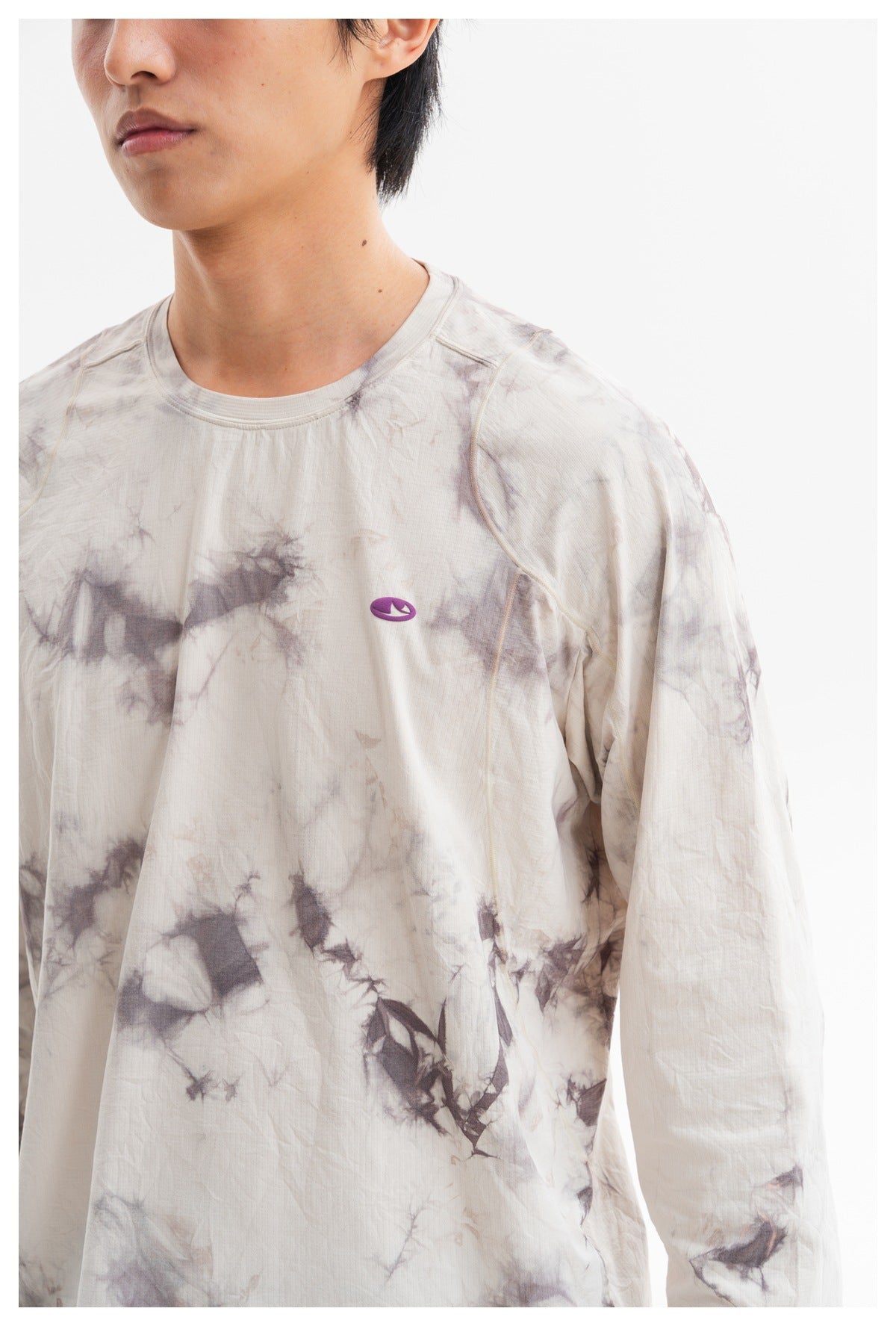 Close-up of the cream and gray tie-dye shirt's front, showing its crew neck, seam details, and small purple chest emblem.