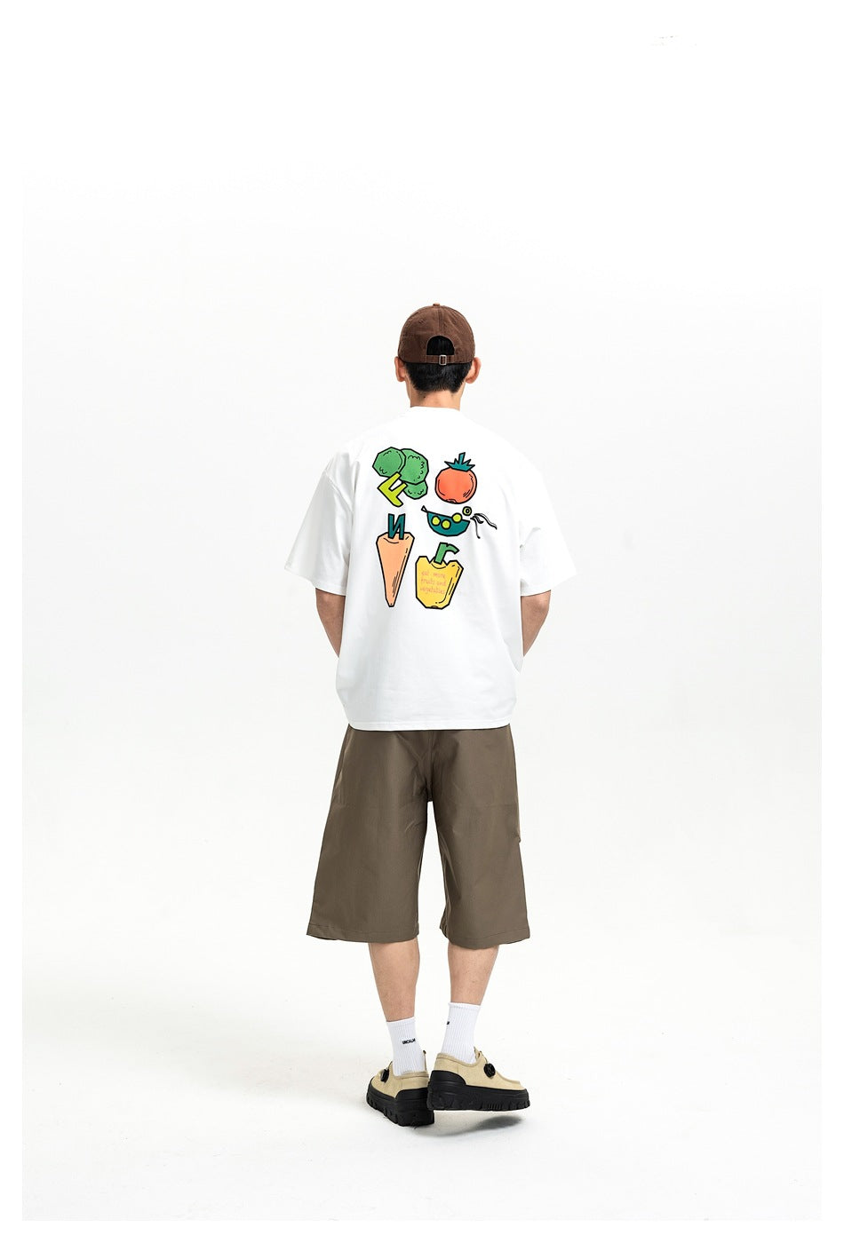 Person seen from behind wearing a white T-shirt with a colorful vegetable print, brown shorts and a brown cap