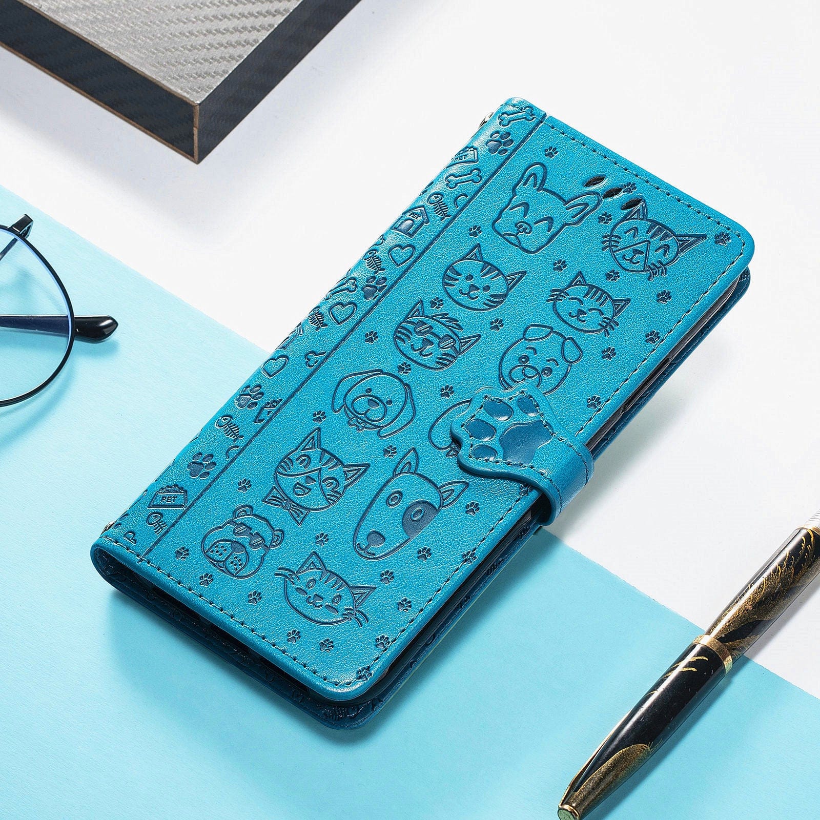 An iPhone 16 Pro Max Wallet Case with Cute Cat and Dog Embossed Design, made from PU leather and featuring card slots and a stand, is placed beside glasses and a pen on a blue and white surface.