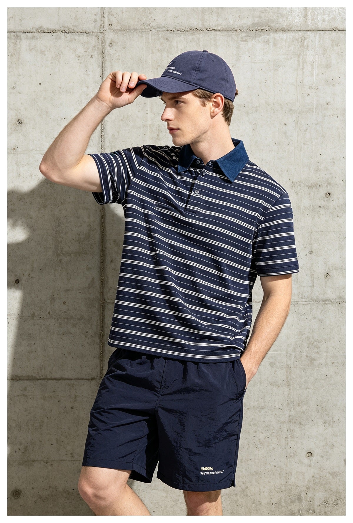 Man in a navy striped polo shirt with blue collar, adjusting his cap against a concrete wall