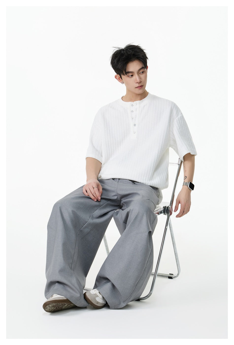 Seated model wearing a white short-sleeve ribbed Henley shirt with a button placket and gray wide-leg trousers.