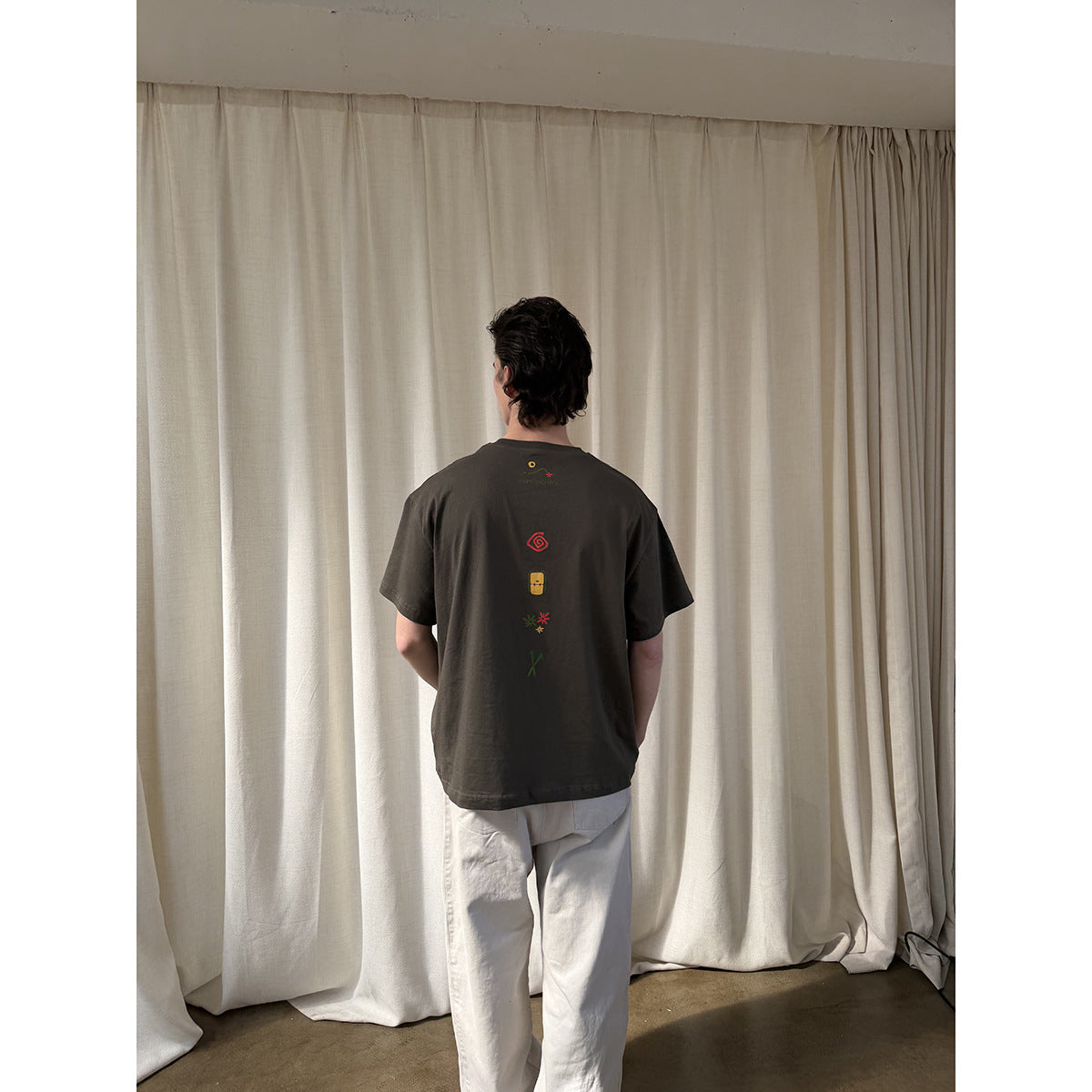 Back view of a person wearing a dark olive T-shirt with a vertical row of small colorful graphics.