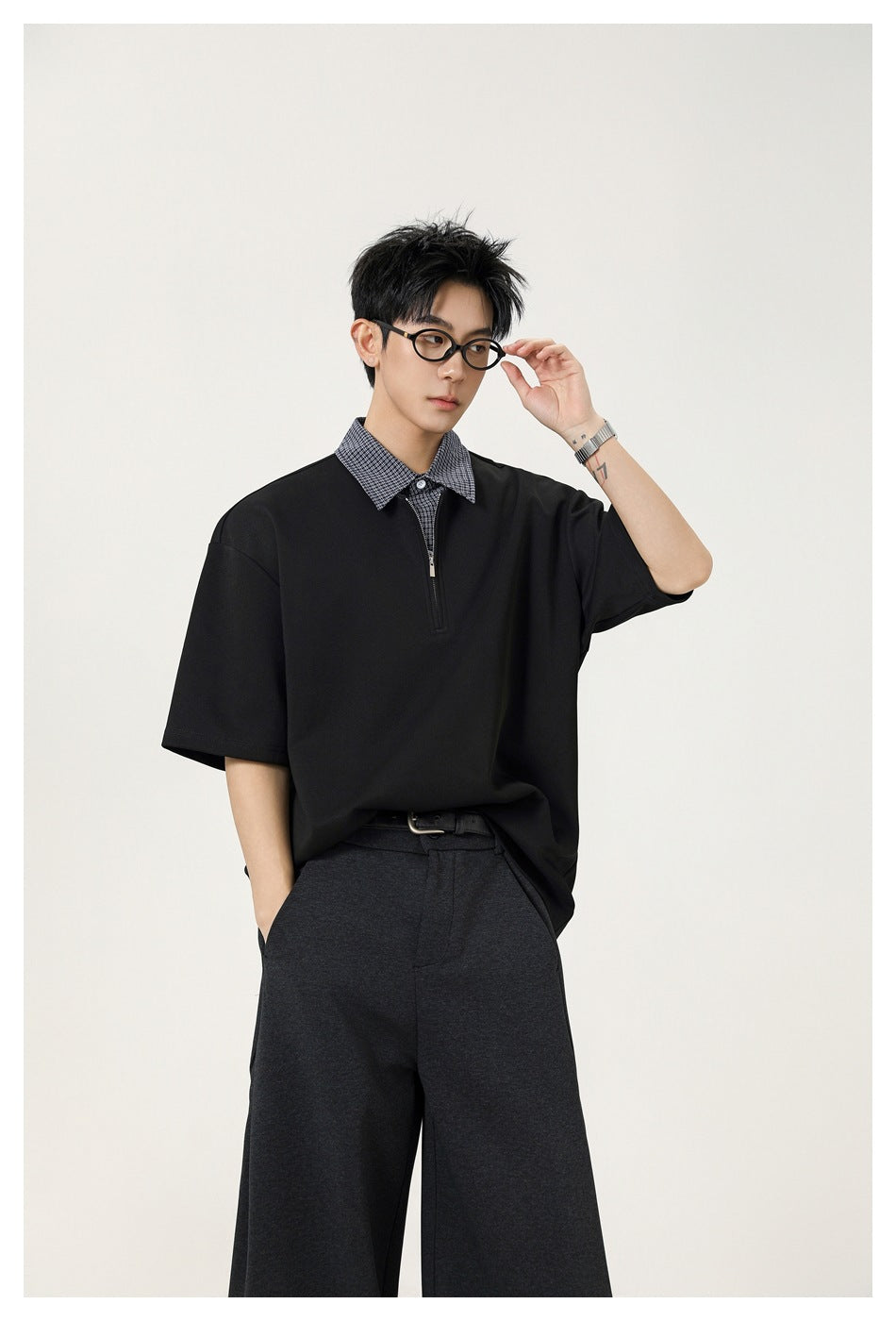 Male model in a black oversized short-sleeve top with a checked collar and short zip placket, adjusting his glasses.