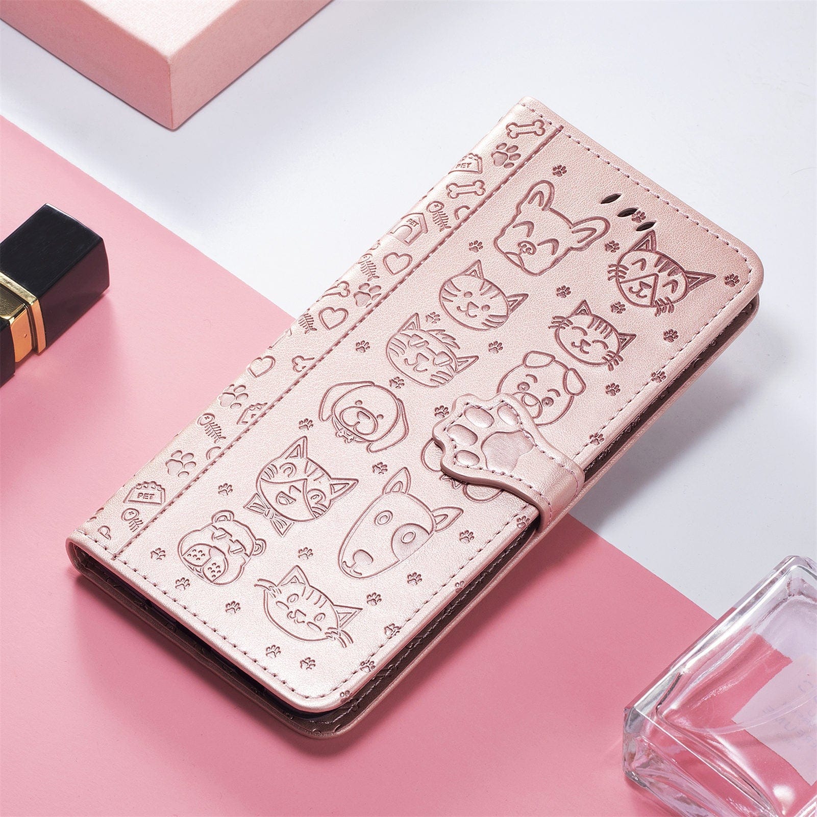 The iPhone 16 Pro Max Wallet Case, featuring a cute cat and dog embossed design on pink PU leather, complete with a paw-shaped clasp, is photographed on a pink surface next to a lipstick and a clear container.