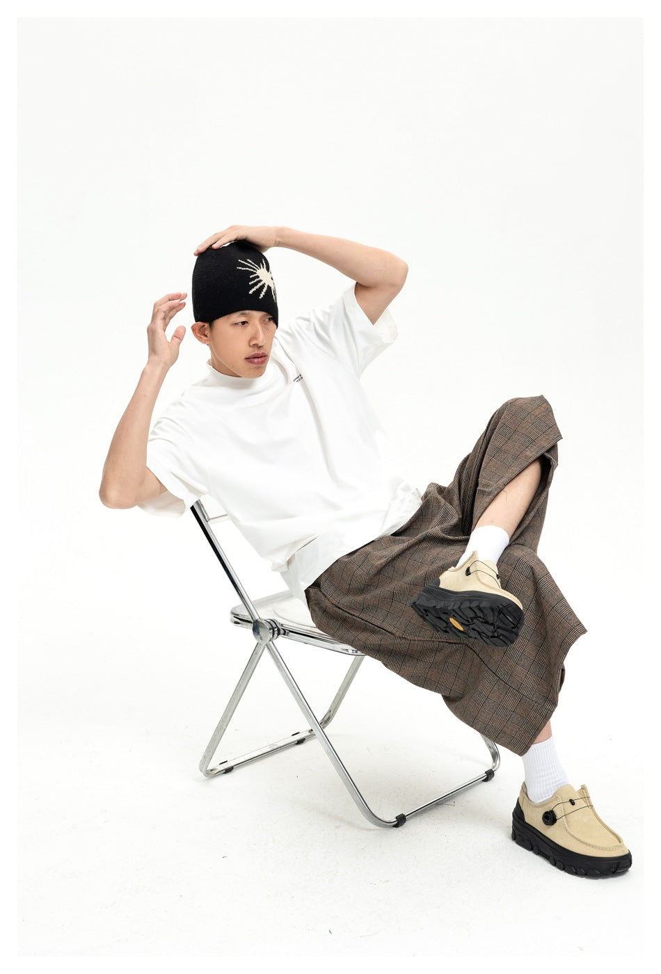 Seated model wearing a white T-shirt, brown plaid wide-leg trousers, black beanie, and beige shoes.