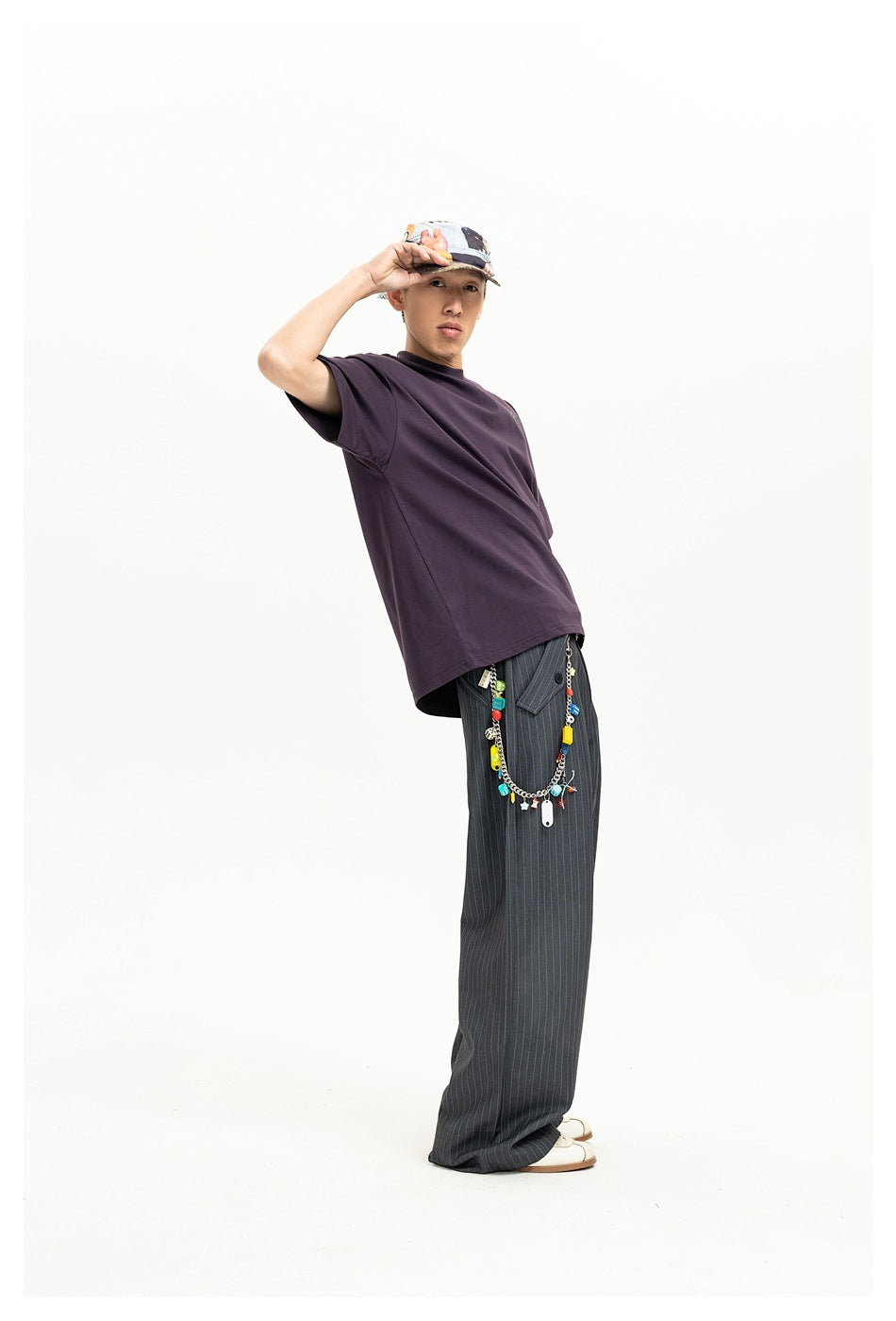Side view of a model wearing a dark plum oversized T-shirt and pinstriped trousers, with a colorful chain accessory at the