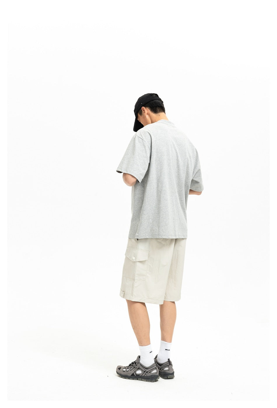 Rear view of a model wearing a plain light gray T-shirt, cream cargo shorts, and a black cap.