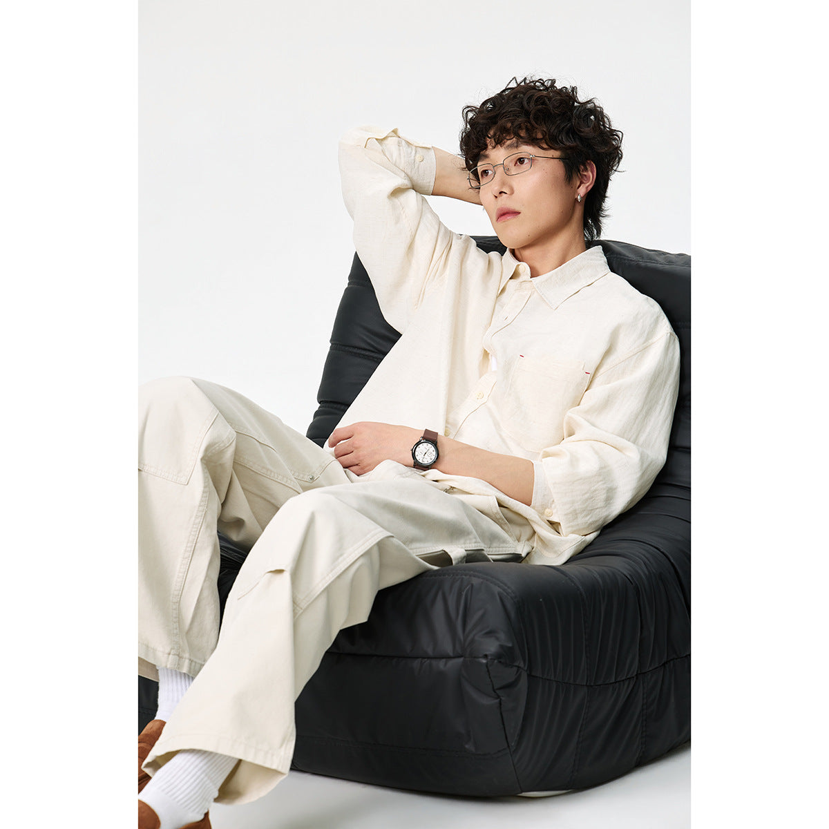 Model seated in a black chair wearing an ivory button-up shirt and matching trousers.