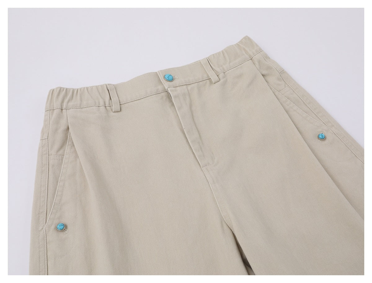 Close-up of beige trousers showing the waistband, belt loops, fly, and turquoise buttons at the sides.