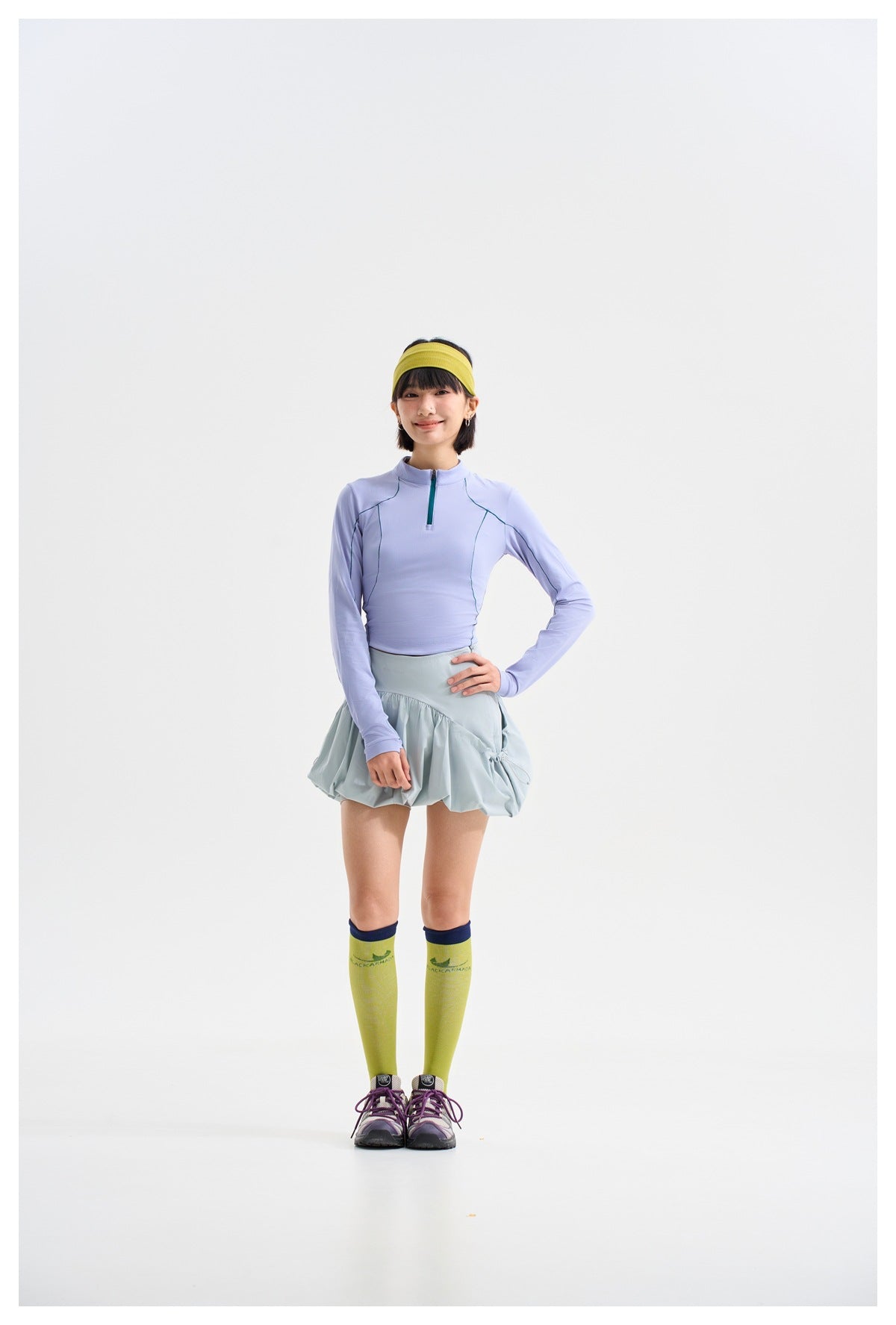 Model wearing a lavender quarter-zip top, pale blue-gray gathered skirt, lime knee-high socks and sneakers, front view.