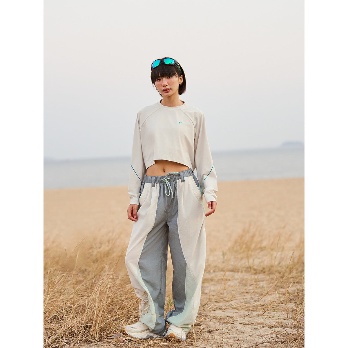 Model wearing a cream cropped long-sleeve top with blue sleeve piping and loose cream-and-gray pants outdoors.